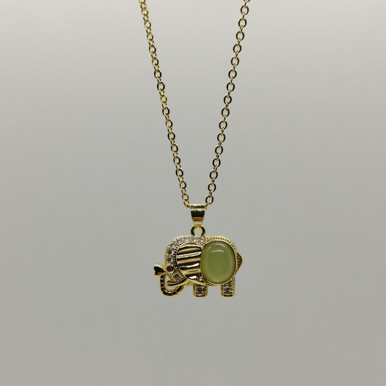 May include: A gold chain necklace with an elephant pendant. The elephant is decorated with clear rhinestones and has a green gemstone in the center.