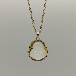 May include: A gold chain necklace with a white jade pendant featuring a carved laughing Buddha design. The pendant is surrounded by a border of small clear rhinestones.