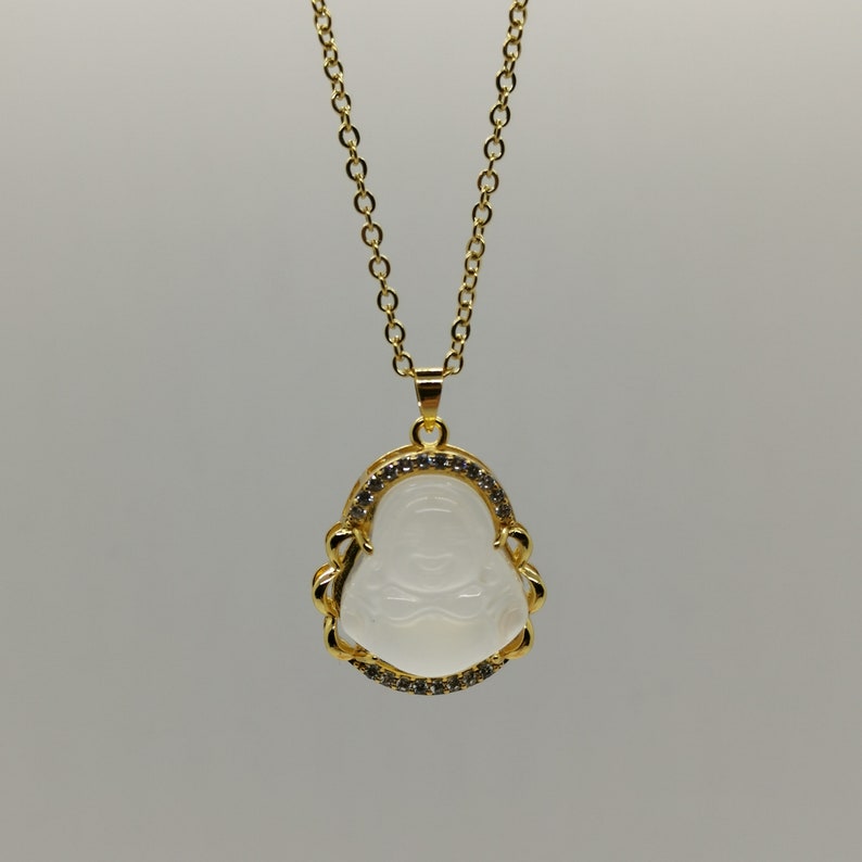 May include: A gold chain necklace with a white jade pendant featuring a carved laughing Buddha design. The pendant is surrounded by a border of small clear rhinestones.