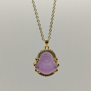 May include: A gold chain necklace with a purple stone pendant featuring a carved image of a smiling Buddha. The pendant is surrounded by a gold frame with clear stones.