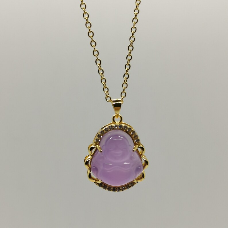 May include: A gold chain necklace with a purple stone pendant featuring a carved image of a smiling Buddha. The pendant is surrounded by a gold frame with clear stones.