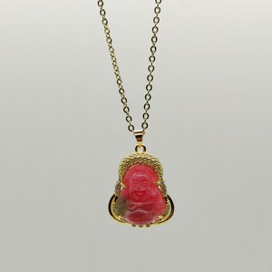 May include: A gold chain necklace with a pink and gold pendant featuring a smiling Buddha. The pendant is adorned with small clear stones.