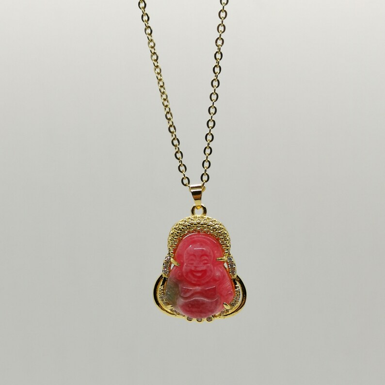 May include: A gold chain necklace with a pink and gold pendant featuring a smiling Buddha. The pendant is adorned with small clear stones.
