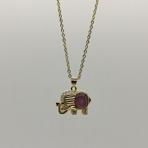 May include: A gold chain necklace with a pendant shaped like an elephant. The elephant is decorated with small clear stones and has a purple stone in its center.