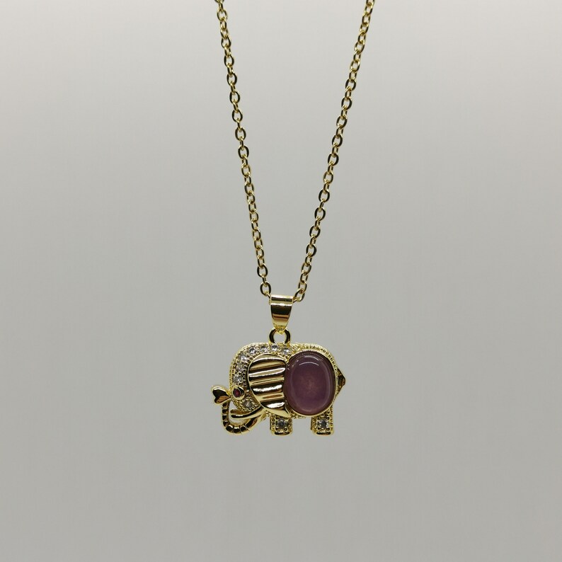 May include: A gold chain necklace with a pendant shaped like an elephant. The elephant is decorated with small clear stones and has a purple stone in its center.