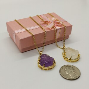 May include: Two gold necklaces with pendants featuring a smiling Buddha face. One pendant is purple and the other is white. The necklaces are displayed in a pink gift box with a gold ribbon.