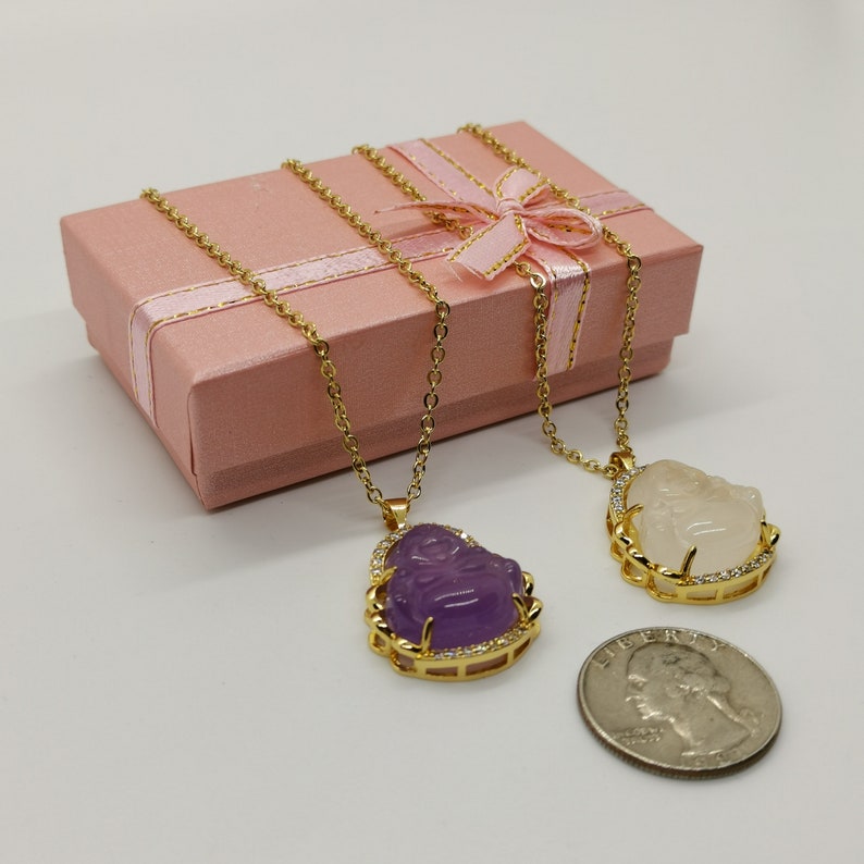 May include: Two gold necklaces with pendants featuring a smiling Buddha face. One pendant is purple and the other is white. The necklaces are displayed in a pink gift box with a gold ribbon.