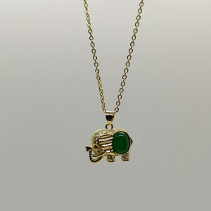 May include: A gold chain necklace with a green gemstone elephant pendant. The elephant has clear rhinestones around the edge and a red stone for the eye.