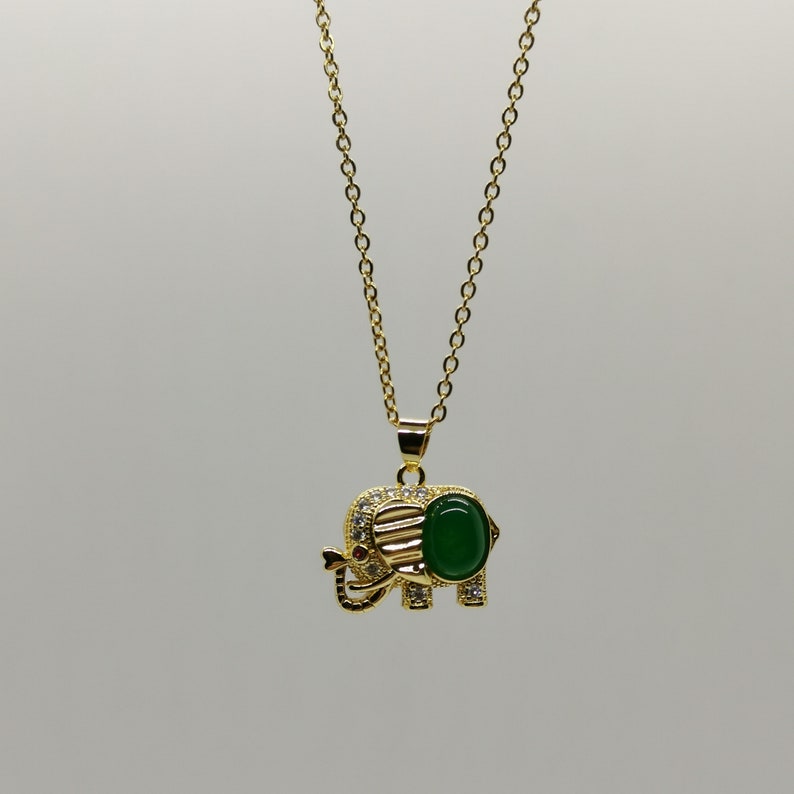 May include: A gold chain necklace with a green gemstone elephant pendant. The elephant has clear rhinestones around the edge and a red stone for the eye.
