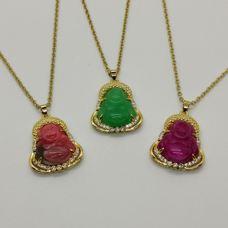 May include: Three gold necklaces with pendants featuring a smiling Buddha figure. The Buddha figures are made of green, pink, and red stone and are surrounded by small clear stones.