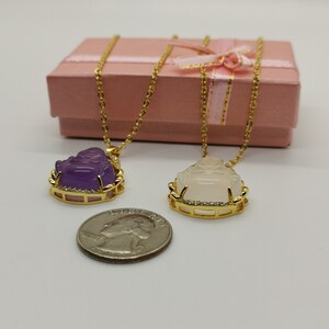 May include: Two gold necklaces with pendants featuring a sitting Buddha figure. One pendant is made of purple jade and the other is white jade. Both pendants are set in a gold frame with clear stones.