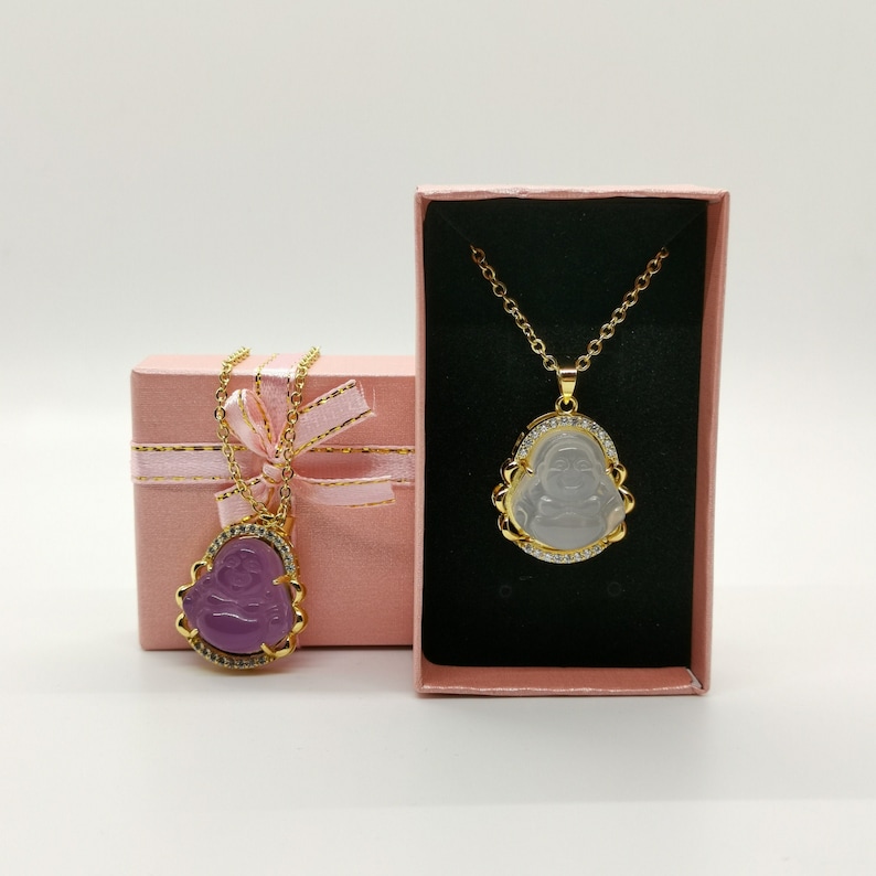 May include: Two gold necklaces with pendants in the shape of a Buddha. One pendant is clear and the other is purple. Both pendants are set with small clear stones. The necklaces are in pink gift boxes.