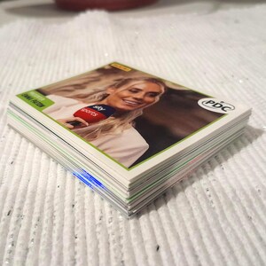 May include: A stack of trading cards featuring a woman holding a microphone with the "Sky Sports" logo. The cards have a green border and the "PDC" logo. The cards are stacked on a white, textured surface.