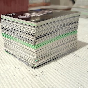 May include: A stack of rectangular cards with a black and white design on the top card. The edges of the cards are white, with some green accents. The stack is on a white, textured surface.