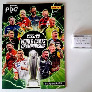 Puede incluir: Un álbum de cromos del Campeonato Mundial de Dardos 2025/26 con jugadores de dardos en la portada. El álbum es verde y negro. También se ve un paquete de 105 cromos.