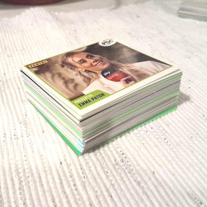 May include: A stack of Panini trading cards featuring a woman with blonde hair. The cards have a green border and the text "sky sports" and "PRESENTER EMMA PATON". The cards are stacked on a white textured surface.