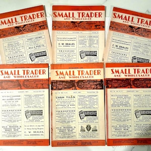 Könnte beinhalten: Eine Sammlung von Vintage-Magazinen "Small Trader and Wholesaler". Jedes Magazin hat ein rot-weißes Cover mit dem Titel und verschiedenen Anzeigen. Die Zeitschriften sind auf einer weißen Oberfläche angeordnet.