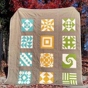 May include: A handmade quilt with a neutral brown background and nine square blocks. Each block features a unique geometric design in shades of white, teal, yellow, and green. The quilt is displayed outdoors with a backdrop of autumn foliage.