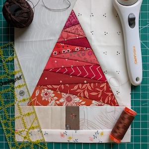 May include: A handmade quilt block featuring a red and white Christmas tree design. The tree is constructed from various red patterned fabrics. A clear ruler, rotary cutter, and spools of brown thread are also present.
