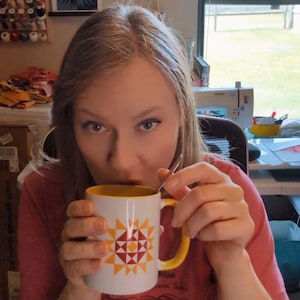 May include: A white mug with a yellow interior and handle, featuring a geometric design in red, yellow, and orange. The mug is held by a person, with a spoon inside. The person is wearing a pink shirt.