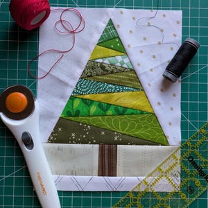 May include: A patchwork Christmas tree quilt block in shades of green. A red reel of thread, a black reel of thread, a rotary cutter, and a clear ruler are also visible. The quilt block is on a green cutting mat.