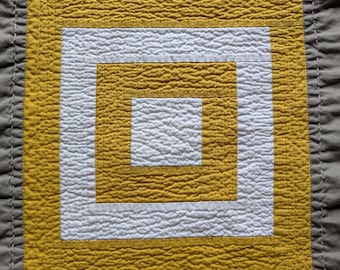 May include: Close-up of a square quilt with a geometric design. The quilt features a central white square surrounded by a mustard yellow square, then another white square, and finally a mustard yellow square. The border is a solid gray.