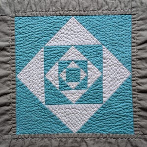 Könnte beinhalten: Quadratischer Quiltblock mit geometrischem Design. Das zentrale Muster zeigt eine Reihe von ineinander verschachtelten Rauten in Weiß und Türkisblau. Der Block wird von einem grauen Rand mit genähten Kanten umrahmt, wodurch ein strukturiertes Aussehen entsteht. Der Quilt ist wahrscheinlich handgefertigt.
