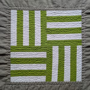 Puede incluir: Textil acolchado cuadrado con un diseño geométrico. El patrón central se compone de rayas verticales y horizontales verdes y blancas alternas, creando un efecto tejido. El diseño está enmarcado en un cuadrado gris, con un borde gris y bordes cosidos.