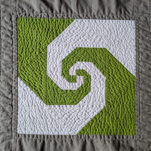 May include: A square quilt block featuring a green and white spiral design. The spiral is centered within a square, with a textured, quilted appearance. The border is a solid gray fabric.