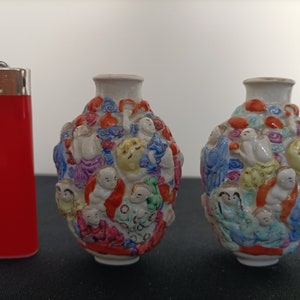 Peut inclure: Deux petits vases en porcelaine blanche avec des motifs complexes de personnages et de fleurs colorés. Les vases sont à peu près de la taille d'un briquet.