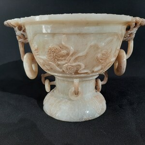 May include: A white jade bowl with intricate carvings of flowers and leaves. The bowl has three handles with ring-shaped ends. The bowl is on a pedestal with a carved base.