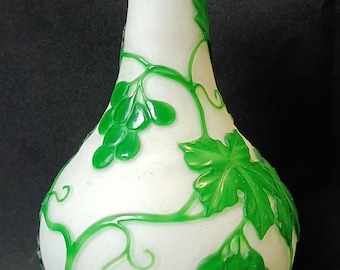 Puede incluir: Un jarrón de vidrio blanco con la parte superior verde y un diseño de vid verde. El jarrón tiene un cuerpo redondeado y un cuello estrecho.