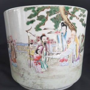 May include: A white ceramic planter with a hand-painted scene of people in a garden. The scene features a large tree, a woman holding a basket of fruit, and several other figures. The planter has a slightly distressed look, with some wear and tear on the surface.