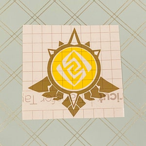 May include: A gold and yellow vinyl decal featuring a stylized sun with a geometric design inside. The decal is on a white gridded background.