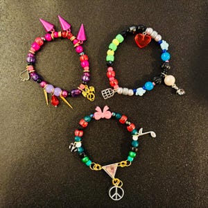 May include: Three beaded bracelets featuring colorful plastic and metallic beads. The first has pink spikes, a spade, and a dragon charm. The second includes a red heart, a birdcage, and blue star beads. The third features a pink bow, a music note, and a peace sign.