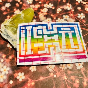 Op de afbeelding: Een kleurrijke sticker met een regenboogverloop en het woord "OPEN" in een pixellettertype. De sticker staat op een rood raster en naast een lichtgroene steen. De achtergrond heeft een bloemenpatroon.