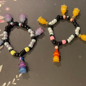 May include: Two colorful beaded bracelets with flower-shaped charms. The bracelet on the left has purple, blue, and yellow beads with a purple, blue, and pink flower charm. The bracelet on the right has pink, white, and black beads with a pink, orange, and yellow flower charm.