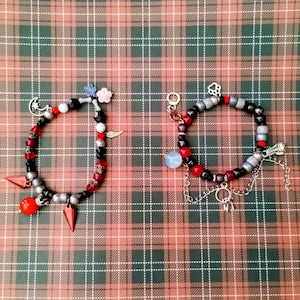 Op de afbeelding: Twee kralenarmbanden met bedels. De armband aan de linkerkant heeft rode, zwarte, zilveren en witte kralen met een halve maan, een rood-wit hart, een roze bloem en een zilveren veerbedel. De armband aan de rechterkant heeft rode, zwarte, zilveren en grijze kralen met een pootprint, een zilveren sleutel, een zilveren ketting en een blauwe glazen kraalbedel.