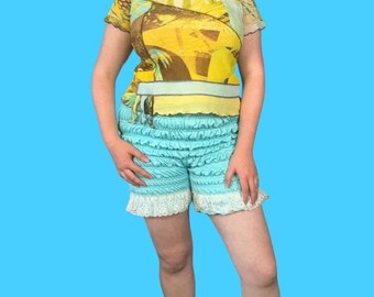 Op de afbeelding: Een vrouw draagt een blauwe en witte gerimpelde short met een geel en bruin geprint doorschijnend topje. De short heeft een witte kantversiering.