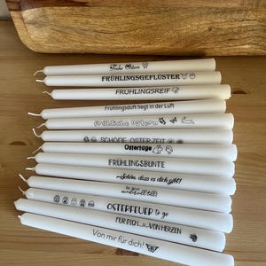 Könnte beinhalten: Eine Ansammlung weißer Stabkerzen mit schwarzem Text und kleinen Illustrationen. Die Kerzen tragen verschiedene Aufschriften wie "Frohe Ostern" und "Frühlingsgeflüster". Die Kerzen liegen auf einer Holzoberfläche.