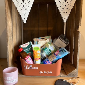 Könnte beinhalten: Eine Holzkiste mit einem dekorativen Papierbanner und einem terrakottafarbenen Behälter, der mit Wellnessprodukten gefüllt ist. Der Behälter trägt die Aufschrift "Wellness" und "für die Seele". Links befindet sich ein rosa Becher mit Text.