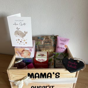 Könnte beinhalten: Eine Holzkiste gefüllt mit Pflegeprodukten, darunter Tee, Hautpflegeprodukte und eine Karte mit einer Walillustration. Auf der Kiste steht "MAMA'S AUSZEIT".