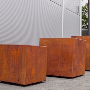 May include: Three square planters made of weathered corten steel with a distinct orange-brown rusted patina. The planters are arranged in a row, each featuring a solid cubic shape with sharp edges and a textured, oxidised metal surface finish.