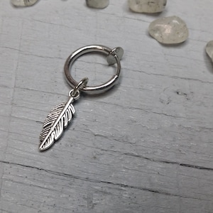 Può includere: Orecchino a clip d'argento con un ciondolo a forma di piuma. L'orecchino è un semplice cerchio con una piccola chiusura argentata. Il ciondolo a forma di piuma è dettagliato e pende dal cerchio.