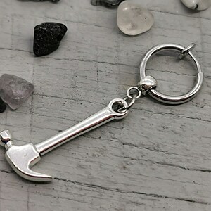 May include: Silver hammer charm attached to a silver hoop earring. The hammer charm has a rounded head and a long handle.
