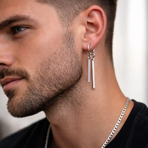 May include: Silver bar earrings hanging from a silver hoop. The earrings are rectangular and dangle. A silver chain necklace is also visible. The person in the image is wearing a black shirt.