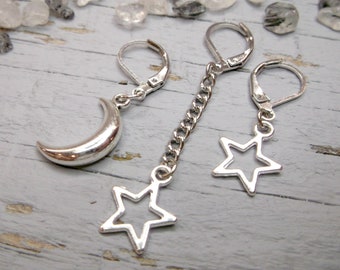 May include: A pair of silver earrings with a crescent moon charm and a star charm hanging from a chain. The earrings are on a white wooden surface.