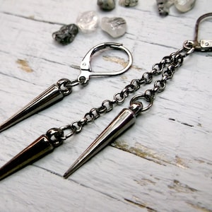 May include: A pair of dark grey, edgy earrings. Each earring features a long, pointed spike pendant suspended from a chain. The earrings have a lever-back closure. Small, clear stones are scattered in the background.