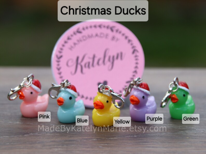 May include: Five Christmas duck charms in pink, blue, yellow, purple, and green. Each duck is wearing a red Santa hat and has a silver clip.