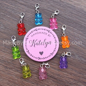 May include: Eight translucent gummy bear charms in red, blue, pink, orange, purple, yellow, light green, and light pink, each with a silver lobster clasp. A circular pink tag in the center displays the text Handmade By Katelyn and an Etsy URL.